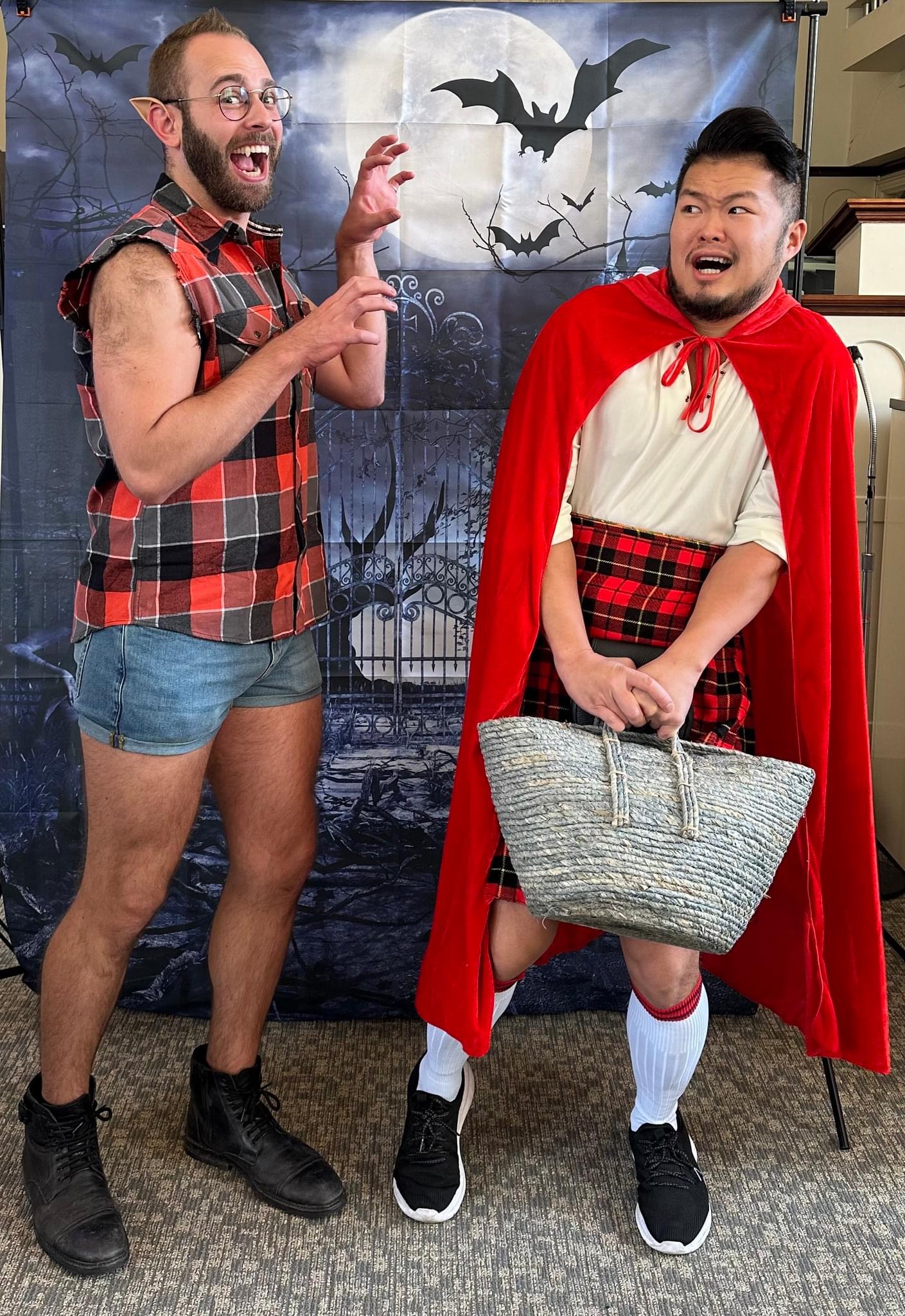 A photo of Michael and Andrew standing in front of a spooky haunted gate backdrop. Michael is dressed as little red riding hood in a red cape and skirt. Andrew is dressed with pointed ear caps and fangs.